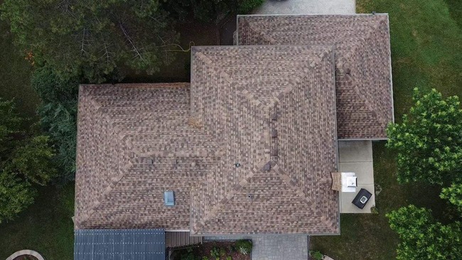 Aerial view of house with uniquely patterned brown and beige asphalt shingle roof, solar panel on lower slope, chimney structure, surrounding trees, and visible driveway.